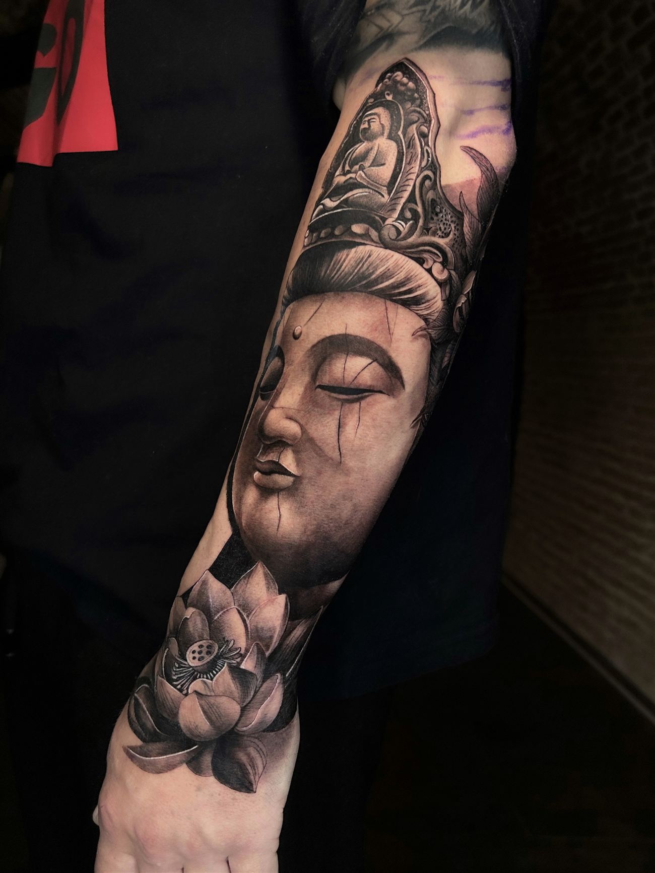 Tatuaje de microrealismo en blanco y negro de una figura de Guanyin sobre un antebrazo, con flor de loto